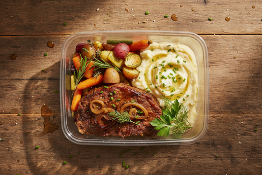 Prepared meatloaf with main , vegetables, and mashed potatoes in a clear container on a wooden surface