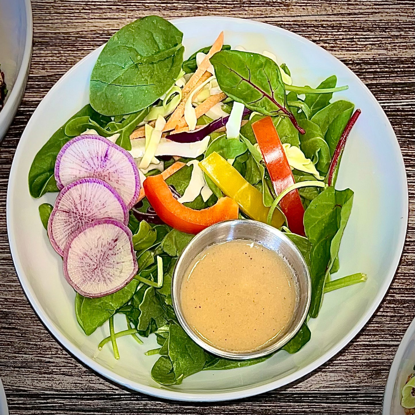 A plate of fresh greens salad with sesame soy vinaigrette, radish slices, and bell pepper strips, accompanied by a small bowl of dressing.