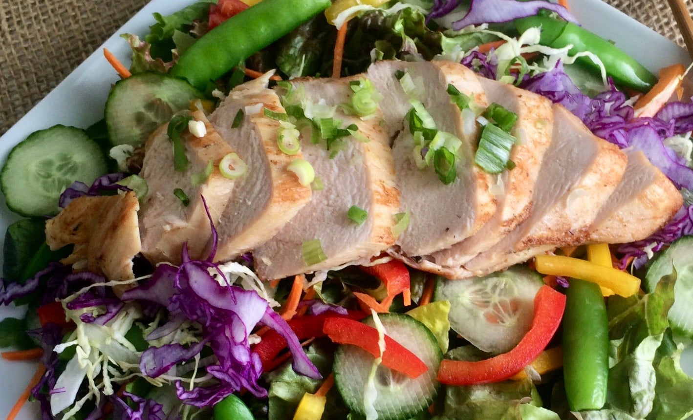 A plated salad featuring sliced cooked meat on a bed of mixed greens, snap peas, cucumber, red and yellow bell peppers, purple cabbage, and lime wedges, served on a white square plate with chopsticks on a burlap surface.